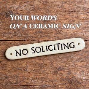 May include: A long, narrow, off-white ceramic sign with rounded ends and two small holes for mounting. The text NO SOLICITING is embossed in black, capital letters across the center of the piece. The surface has a smooth, glossy finish.
