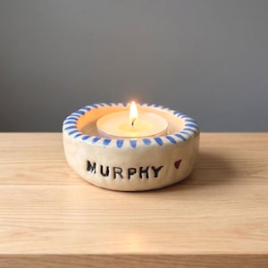 May include: A round, off-white ceramic candle holder with a blue-striped rim. The side features the black text MURPHY followed by a small red heart. A lit white tea light candle sits in the centre of the bowl, casting a warm, glowing light.