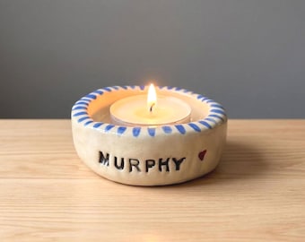 May include: A round, off-white ceramic candle holder with a blue-striped rim. The side features the black text MURPHY followed by a small red heart. A lit white tea light candle sits in the center of the bowl, casting a warm, glowing light.
