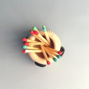 May include: A small, round, cream-coloured ceramic holder containing a cluster of wooden matches. The match heads are painted in various colours, including red, light green, and white. The holder features a smooth, rounded rim and dark accents.