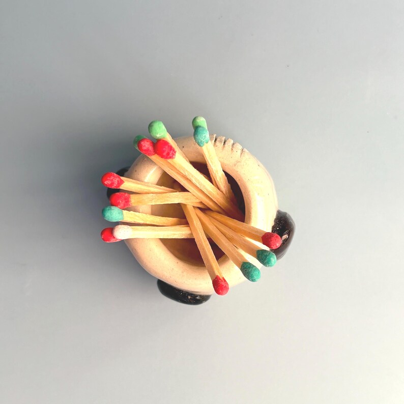 May include: A small, round, cream-coloured ceramic holder containing a cluster of wooden matches. The match heads are painted in various colours, including red, light green, and white. The holder features a smooth, rounded rim and dark accents.