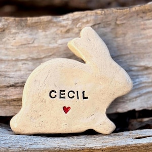 May include: A beige ceramic rabbit figurine with a smooth, glossy finish. The name CECIL is stamped in black capital letters on the side of the rabbit, positioned above a small, painted red heart. The rabbit is depicted in a seated profile.