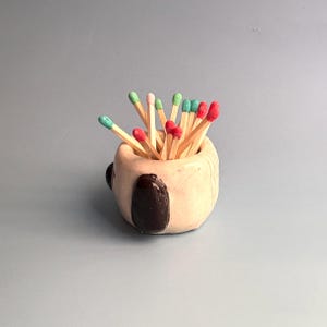 May include: A small, round, beige ceramic holder with a glossy finish and dark brown accents, filled with wooden matches that have vibrant red and green tips. The container has a smooth, hand-formed appearance with visible texture and depth.