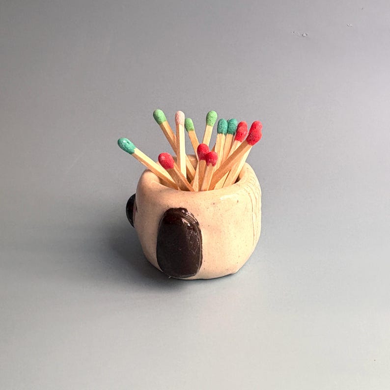 May include: A small, round, beige ceramic holder with a glossy finish and dark brown accents, filled with wooden matches that have vibrant red and green tips. The container has a smooth, hand-formed appearance with visible texture and depth.