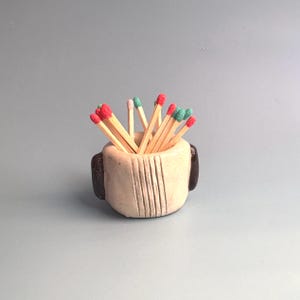 May include: A small, round, beige ceramic holder with dark brown side accents and vertical line textures, filled with wooden matches featuring red, pink, and green tips. The container has a thick, hand-formed appearance with a smooth finish.