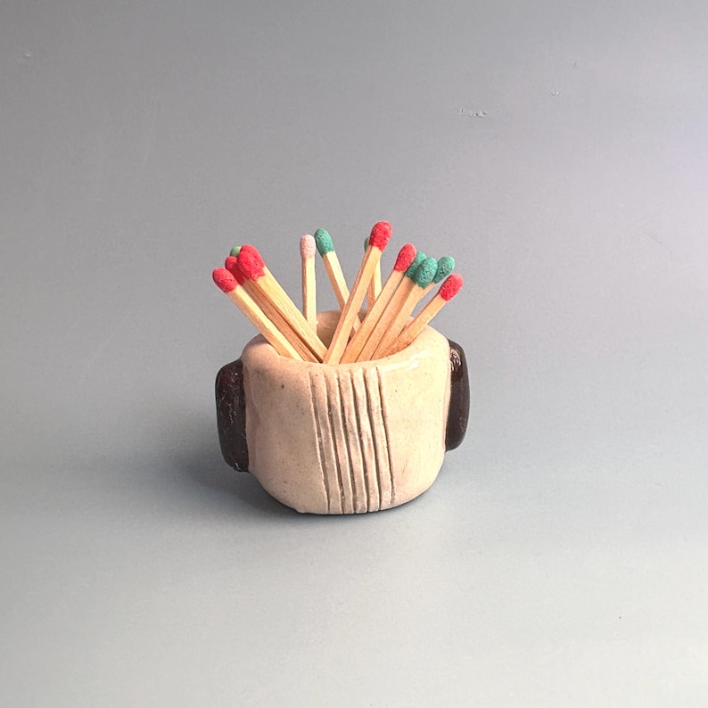 May include: A small, round, beige ceramic holder with dark brown side accents and vertical line textures, filled with wooden matches featuring red, pink, and green tips. The container has a thick, hand-formed appearance with a smooth finish.