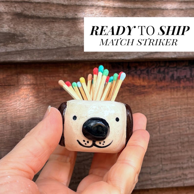 May include: A small, cream-coloured ceramic match striker shaped like a dog head with dark brown ears, a black nose, and painted eyes. The container is filled with wooden matches featuring colourful tips in shades of red, blue, green, and yellow.