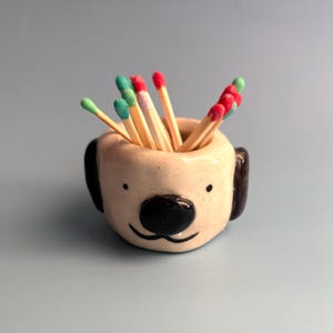 May include: A small, beige ceramic holder shaped like a dog's head, featuring dark brown ears, a black nose, and two small black eyes. The holder is filled with several wooden matches that have colourful red, green, and white tips.