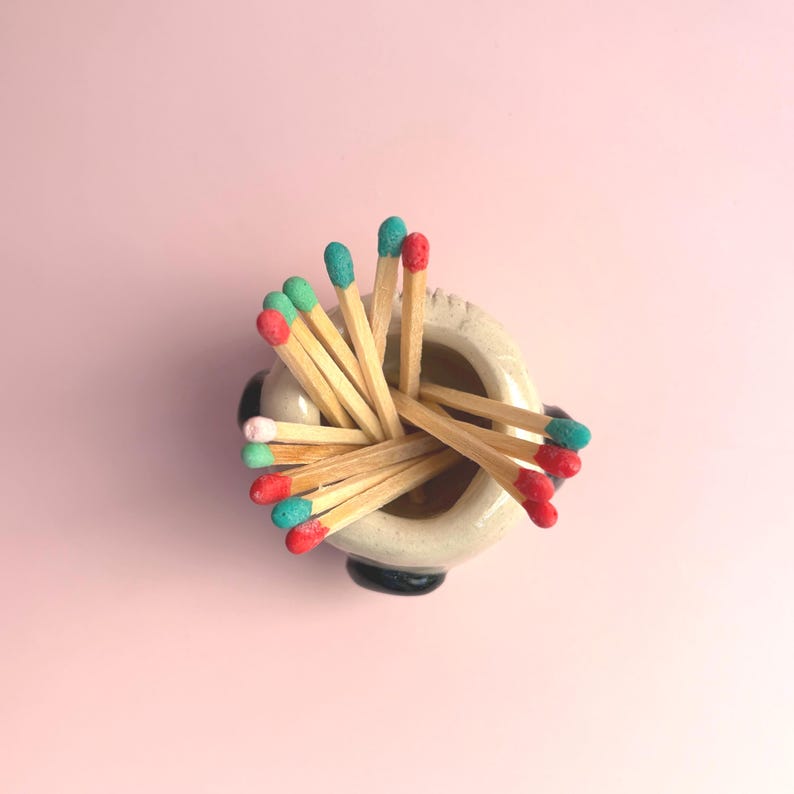 May include: A small, round, off-white ceramic holder containing a collection of wooden matches. The match heads are painted in various colours, including bright red, teal, and soft pink, arranged in a cluster pointing upward from the vessel.