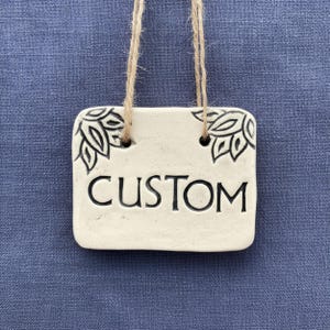 May include: A rectangular, off-white ceramic sign with the word "CUSTOM" in black, centered. The sign has decorative leaf designs in black at the top corners and is suspended by a natural twine string. The background is a textured blue fabric.