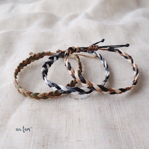 May include: Three braided friendship bracelets in brown, black, and white. The bracelets are tied together and laid out on a beige fabric background. The text "an tam bracelets" is visible in the image.