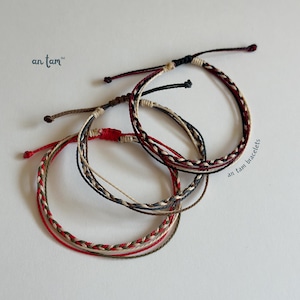 May include: Three braided bracelets in red, black, brown, and beige. The bracelets are made of woven cord and are displayed on a white surface. The bracelets have the text "an tam bracelets" and the brand name "an tam" on them.