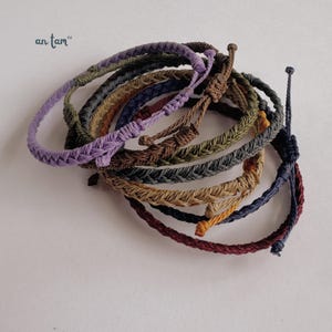 May include: A selection of braided rope bracelets in shades of purple, green, brown, and blue. The bracelets are knotted and appear handmade. The brand name "an tam" is visible in the upper left corner.