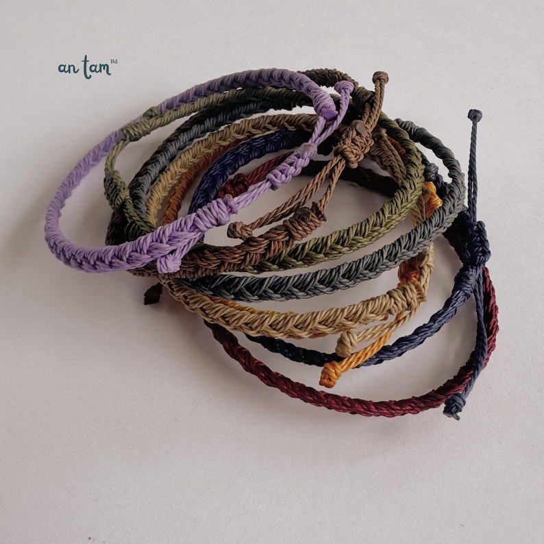 May include: A selection of braided rope bracelets in shades of purple, green, brown, and blue. The bracelets are knotted and appear handmade. The brand name "an tam" is visible in the upper left corner.