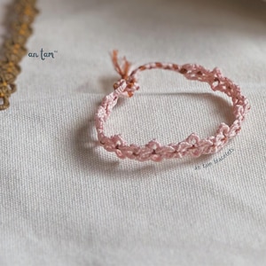 May include: A pink braided friendship bracelet with a knotted design. The bracelet is on a white fabric background. The text "an tam bracelets" is visible in the lower right corner of the image.