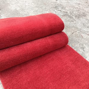 Può includere: Un tappeto rosso brillante arrotolato è appoggiato su una superficie di cemento. Il tappeto è realizzato in un materiale strutturato, con un pelo visibile. La sezione arrotolata mostra lo spessore del tappeto, e la parte srotolata si estende verso il basso.