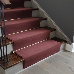 Op de afbeelding: Een trap met bordeauxrode tapijtlopers en donkere houten treden. De trap heeft een wit geschilderde basis en een zwarte metalen balustrade. De vloer is lichtgrijs hout.