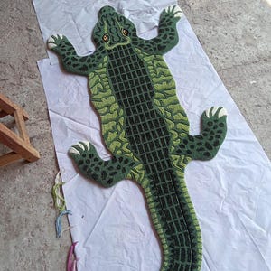 May include: A tufted rug shaped like a crocodile, featuring a dark green and lime green textured pattern with a black grid design along the centre of the back and tail. The rug has four limbs with white claws and two yellow eyes on the head.