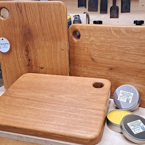 Può includere: Tre taglieri in legno con manico, realizzati in legno di quercia massiccio. I taglieri sono di dimensioni diverse e hanno una finitura in legno naturale. I taglieri sono appoggiati su una superficie in legno con due piccole lattine di balsamo per legno.