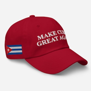 Può includere: Un cappellino da baseball rosso con la scritta "MAKE CUBA GREAT AGAIN" ricamata in bianco. La bandiera cubana è ricamata sul lato. Il cappellino ha una visiera curva e una corona arrotondata.