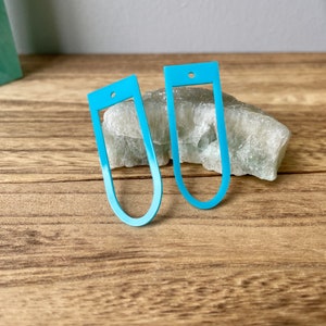May include: Two turquoise blue metal earrings in the shape of an oval with a small hole at the top. The earrings are on a wooden surface.