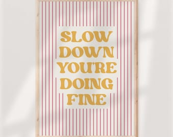 May include: A rectangular framed wall art print featuring a cream-colored background with vertical thin red stripes. Centered in the print is the yellow text that reads: SLOW DOWN YOU'RE DOING FINE, written in a bold, rounded, retro-style typeface.