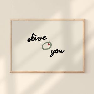 May include: A framed print with the words "olive you" in black script. The "o" in "olive" is replaced with a green olive with a red heart pit. The background is a light beige color, and the frame is a light wood color.