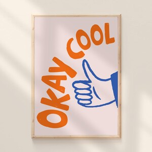 May include: A framed art print with the words "OKAY COOL" in orange, with a blue hand giving a thumbs up. The background is a light pink color, and the frame is a light wood color.