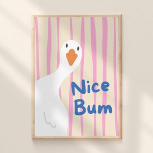 May include: A framed art print featuring a cartoon goose with an orange beak and the words "Nice Bum" in blue. The background has vertical pink stripes on a beige backdrop. The artwork is in a wooden frame.