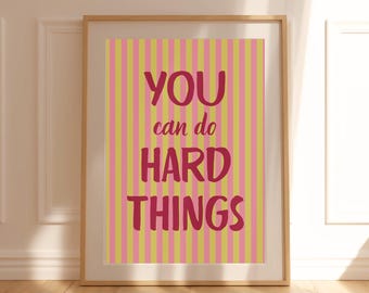 May include: A framed wall art print featuring vertical stripes in alternating shades of yellow and pink. The text "YOU can do HARD THINGS" is printed in a bold, dark red font across the center of the striped background within a light wood frame.