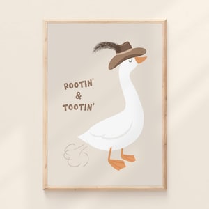 May include: A framed print featuring a whimsical illustration of a white goose wearing a brown hat with a feather. The goose is depicted with orange feet and beak, and the text "ROOTIN' & TOOTIN'" is displayed on the print. The background is a light beige color.