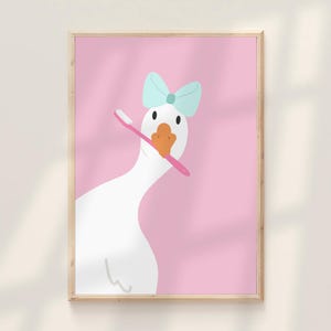 May include: A framed print showing a cartoon goose with a light blue bow and a pink toothbrush. The goose has a white body, orange beak, and black eyes, set against a pink backdrop.