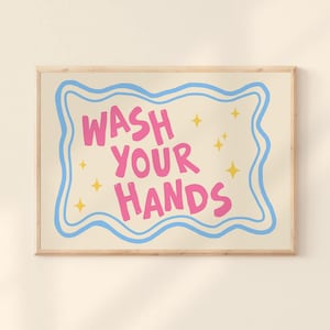 Puede incluir: Un cuadro enmarcado con las palabras "WASH YOUR HANDS" en rosa, delineadas en azul, sobre un fondo crema. Acentos de estrellas amarillas adornan el texto. El marco es de color madera clara.