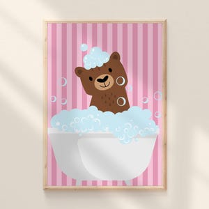 以下が含まれることがあります： 白いバスタブで青い泡に浸かっている茶色のクマの額装イラスト。クマは笑顔で、頭に泡がついています。背景はピンクの縦縞模様です。アートワークは木製のフレームに入っています。