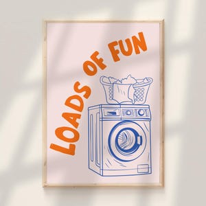 May include: A framed print with a light pink background features a blue and white illustration of a washing machine with two laundry baskets on top. The words "LOADS OF FUN" are in orange.