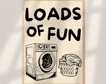May include: A framed print with the words "LOADS OF FUN" in large black letters. Below the text is a black and white illustration of a washing machine and a laundry basket filled with clothes. The background is a light beige color.