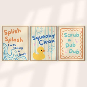 Puede incluir: Juego de tres láminas enmarcadas para el baño. Las láminas presentan textos y gráficos lúdicos en naranja, azul y amarillo. Una lámina dice "Splish Splash" con líneas onduladas, otra "Squeaky Clean" con un patito de goma amarillo y la tercera "Scrub a Dub Dub".