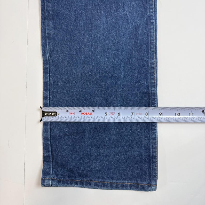 May include: Close-up of blue denim jeans with a measuring tape showing a width of approximately 17.8 cm. The jeans are on a white surface. The tape measure is labeled "Kobalt".