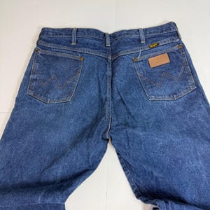 May include: Blue denim jeans with the Wrangler logo on the back pocket and a brown leather patch. The jeans have a classic five-pocket design and a straight leg cut. The denim has a faded, worn look.