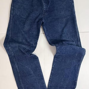 May include: A pair of blue denim jeans, displayed flat on a white surface. The jeans feature a classic five-pocket design, a button and zip closure, and a faded, worn look. The denim is a medium wash, with subtle colour variations.
