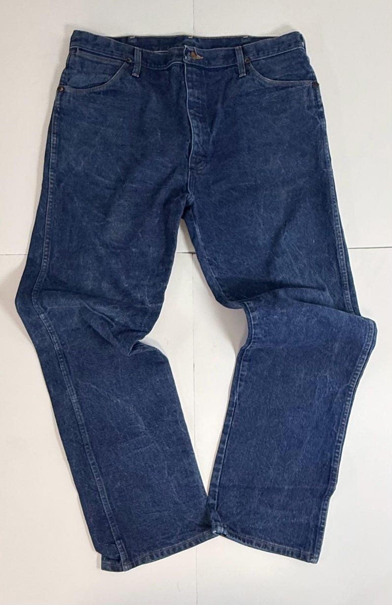May include: A pair of blue denim jeans, displayed flat on a white surface. The jeans feature a classic five-pocket design, a button and zip closure, and a faded, worn look. The denim is a medium wash, with subtle colour variations.