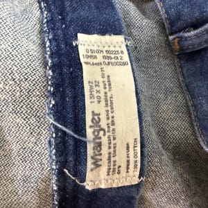 May include: Close-up of a blue denim Wrangler jeans label. The label shows the brand name, size 40 x 32, and care instructions. The jeans are made of 100% cotton.