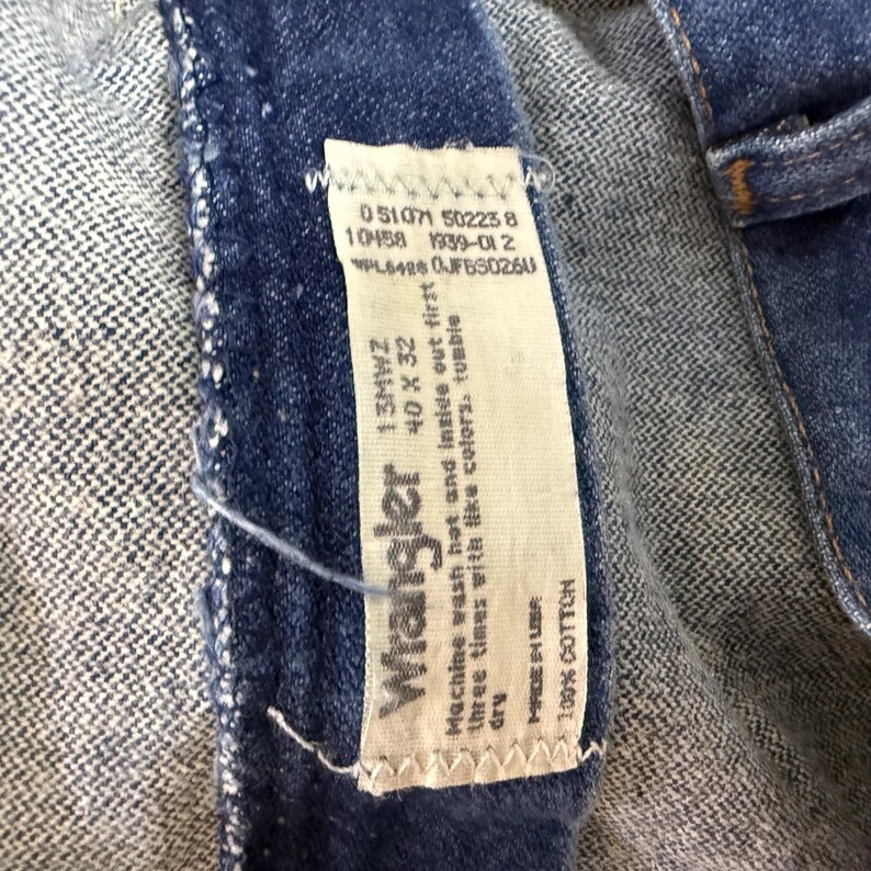 May include: Close-up of a blue denim Wrangler jeans label. The label shows the brand name, size 40 x 32, and care instructions. The jeans are made of 100% cotton.