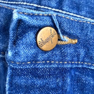 May include: Close-up of blue denim fabric with a brass button. The button is round with a detailed design. The denim has visible stitching and a buttonhole. The overall colour is a medium blue.