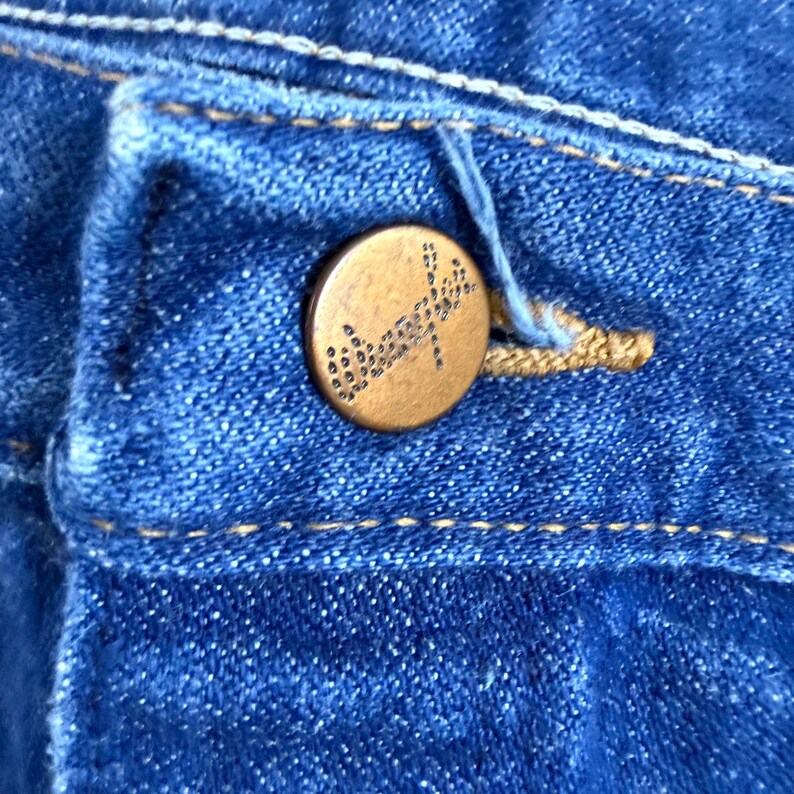 May include: Close-up of blue denim fabric with a brass button. The button is round with a detailed design. The denim has visible stitching and a buttonhole. The overall colour is a medium blue.