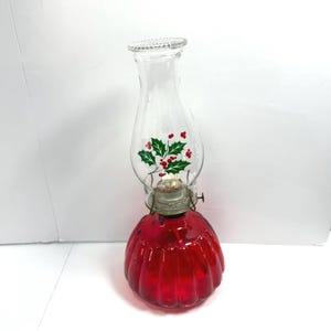 May include: A vintage oil lamp with a clear glass chimney decorated with a holly and berry design. The lamp's base is a vibrant red, and the metal components have a silver finish. The lamp is designed for decorative use.