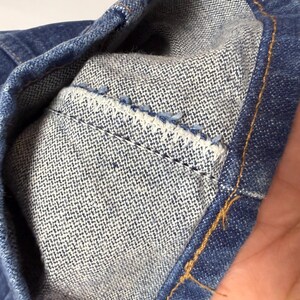 May include: Close-up of denim fabric showing the inside of a pocket. The pocket is made of a lighter blue fabric and is stitched to the darker blue denim. The stitching is visible, and the edges are frayed.