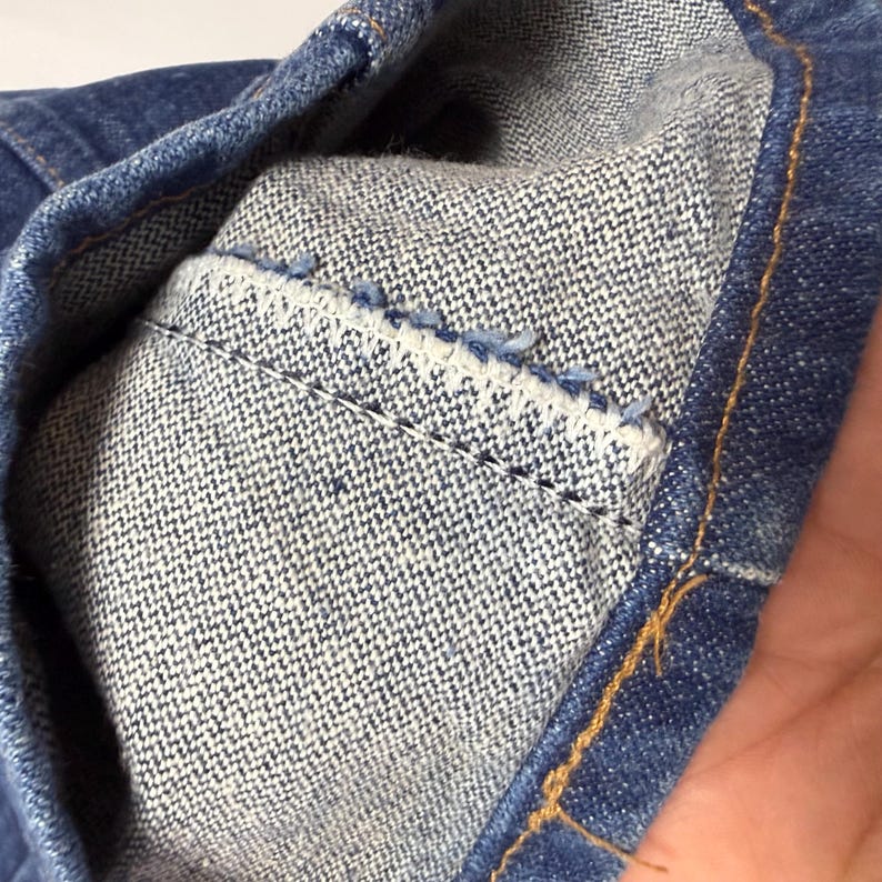May include: Close-up of denim fabric showing the inside of a pocket. The pocket is made of a lighter blue fabric and is stitched to the darker blue denim. The stitching is visible, and the edges are frayed.
