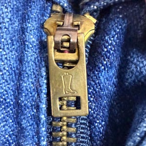 May include: Close-up of a brass zip on blue denim fabric. The zip pull features a boot design. The zip is partially closed, showing the interlocking teeth and the texture of the denim.
