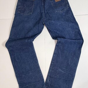 May include: Dark blue denim jeans with a classic five-pocket design. The jeans have a straight leg cut and a brown leather patch on the back pocket. The Wrangler logo is visible on the patch. The jeans are laid flat on a white surface.